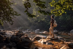 A woman explores new, magical, and fantastic places around the world, surrounded by nature and spreading her arms to breathe and relax. Female hiker crossing the forest creek.