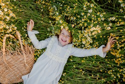 happy little girl in a cotton dress lies in a field of daisies in the summer at sunset. laughs, view from above