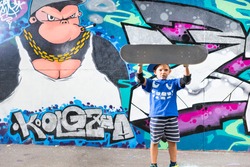 Full Length Portrait of Young Boy Holding Skateboard Above Head in front of Graffiti Covered Wall in Urban Skate Park