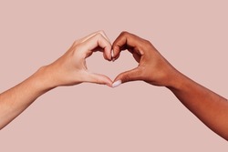 Close up of black and white female hands in heart shape, interracial friendship. Multiracial hands over beige background at studio. Peace and love concept.