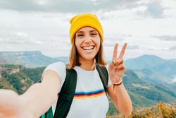 Young happy smiling woman backpacker with short brunette hair and yellow beanie, taking selfie with smartphone doing victory sign with hand on top rocky mountains. Adventure, travel, holiday concept.