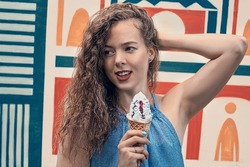 A fair haired curly girl holds an big ice cream and smiles joyfully in a blue tunic against the background of a colorful wall