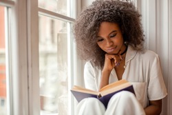 Pretty african american girl with reading a book sitting on the windowsill.