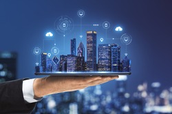Smart city concept with real skyscrapers layout with glowing digital cloud technology icons on digital tablet screen that carrying businessman hand on blurry megapolis city background