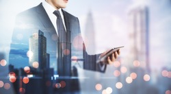 Freelance mobile working concept with man in black suit looking at digital tablet on blurry megapolis city background. Double exposure