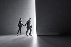 Side view of businessman and woman holding hands and stepping into the light from behind a concrete wall. Success, teamwork and future concept