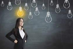 Portrait of attractive thoughtful young european businesswoman standing on chalkboard wall background with glowing drawn light bulb sketch and mock up place. Idea and creativity concept