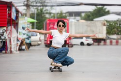 Cheerful Asian young woman skater riding on skateboard on courtyard at gas station with wear sunglasses. Girl skateboarding outdoor