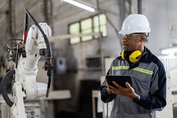 African American male engineer worker using digital tablet control automatic robotic hand machine in factory. Black male technician worker working with control automatic robot arm system welding