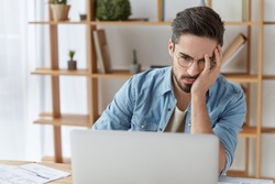 Exhausted embarrassed male reads some information on laptop computer, being tired and overworked after hard work at office all day long, doesn`t understand how make project, being frustrated