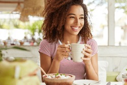 Photo of positive dark skinned mixed race female ejoys good rest at coffee shop, drinks hot beverage, has broad smile, happy to discuss something funny with friends. People, leisure and eating concept