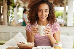 Portrait of good looking African American female drinks hot coffee in restaurant during weekend, smiles happily, enjoys spare time and tasty beverage. Adorable dark skinned woman poses indoor with cup