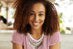 Portrait of happy of dark skinned female with curly bushy Afro hairstyle, smiles broadly into camera, rests in cafe with boyfriennd, being glad to have much spare time. People, ethnicity concept