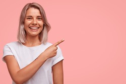 Horizontal portrait of beautiful cheerful Caucasian female with braces, has broad smile, indicates at blank copy space for your promotional content, smiles joyfully. People, advertisement concept