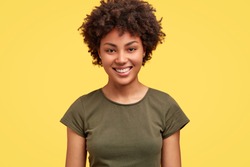 Cheerful African American female with pleasant smile, has curly dark hair, healthy skin, wears casual t shirt, stands against yellow background, being in good mood as husband came from work early