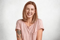 Happy European female laughs joyfully, shows white teeth, has bobbed hairstyle, dressed in casual t shirt, has tattoo, isolated over white background, recieves pleasant compliment. Emotions.