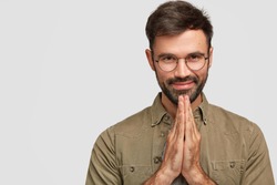 People and meditation concept. Faithful pleased young European man with beard and mustache, keeps palms together, believes in something good, dressed in fashionable shirt, poses indoor alone