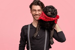 Cheerful bearded male has toothy smile, glad to pose at camera with his pedigree dog, likes pets, dressed in fashionable black shirt and suspenders, isolated over pink wall. Happy man with animal