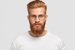 Headshot of attractive serious Caucasian male with thick ginger beard and trendy haircut, dressed in casual white t shirt, looks directly at camera, wears round glasses, isolated over white background