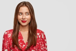 Attractive young European female with long dark hair, red painted lips and stylish blouse, looks pensively aside, has dreamy expression, stands against white background with copy space on left side
