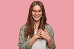 People, dedication and promise concept. Happy girlfriend keeps hands on heart, swears be loyal to boyfriend, expresses gratitude, wears spectacles, casual shirt, stands against pink studio wall.