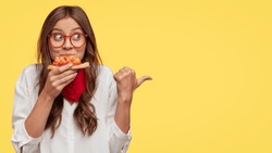 Photo of attractive woman eats slice of pizza, points aside with thumb, dressed in fashionable clothes, shows where pizzeria is, isolated over yellow background. Pretty girl has snack with fastfood