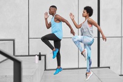 Motivated active ethnic couple run up stairs together, jump highly, train climbing staircase in city, wear comfortable sportsclothes, drink water from bottle, climb challenge, choose difficult path