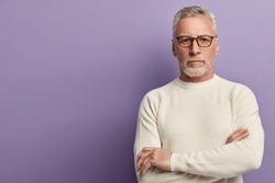 Indoor shot of self confident man with thick stubble, keeps hands crossed, wears transparent spectacles and white sweater, looks directly at camera, isolated over purple background with empty space