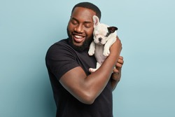 Sweet moments with domestic animals. Portrait of satisfied dark skinned man holds cute small french dog puppy closely to face, enjoys good time with loyal pet, wears casual black t shirt, pose indoor