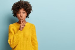 Studio shot of quiet dark skinned lady keeps fore finger over lips, makes silence gesture, gossips with friend, wears yellow sweater, says hush, isolated over blue studio wall with blank space