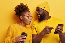 Surprised curious dark skinned woman looks at screen of boyfriends cellular, reads online post with interesting content, excited with smartphone or app features, isolated over yellow background.