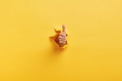 Image of unrecognizable man makes thumb up gesture, demonstrates approval or agreement, gestures through torn paper wall yellow background. Body language concept. Hand sign. Hole in wall. Like gesture