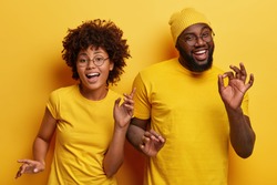 Photo of happy African couple dance together against yellow background, move body actively, show okay gesture, wear casual yellow t shirt, have fun during party. Monochrome. Feeling rhytm of music