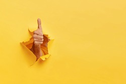 Unrecognizable man shows like gesture through torn yellow wall, keeps thumb up, says you are best, demonstrates approval sign, recommends something. Copy space aside for your advertising content
