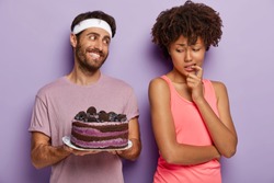 African American woman keeps finger on mouth, feels temptation while looks at delicious blueberry cake, tries not to eat unhealthy food, being on diet and goes in for sport. Mmm, how tasty it looks