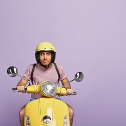 Confident serious man rides on motorcycle, focused aside, stops on way to have rest, wears helmet, casual purple t shirt, covers distance, travels alone, enjoys speed. Professional male rider on bike