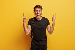 Joyous young hipster listens rock music in stereo headphones, shows heavy metal gesture, keeps mouth opened, dressed in black outfit, models against yellow background. Body language concept.