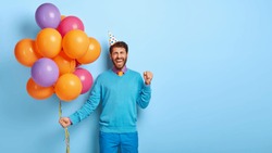 Joyous optimistic man celebrates successful job position, being on corporate party, clenches fist in triumph, holds bunch of colorful balloons, wears blue outfit, party hat, stands indoor, copy space