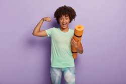 Funny dark skinned woman raises arm, shows muscles after training, holds karemat, has regular workout in gym with coach, dressed in sportswear, isolated on purple studio wall. Strength concept