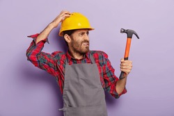 Photo of discontent handyman turns right, looks into distance with frowned face, holds hammer, being professional builder, notices new object for repairing wears helmet, apron. Renovation, engineering