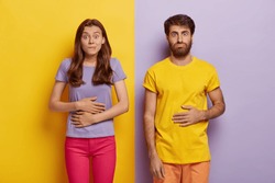 Young female and male touch stomaches, feel satiety after eating hearty meal, bites lips, have good appetite, dressed in casual outfit, pose against yellow and purple wall. Pleasant feeling in belly