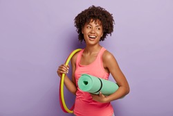 Sporty slim lady with healthy dark skin, Afro hairstyle, exercises with hula hoop, carries rolled up karemat, dressed in pink vest, has toothy smile, poses indoor. Healthy lifestyle, gymnastic concept