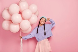 Horizontal view of happy young Asian woman with two pigtails, dreams about awesome holiday, carries bunch of air balloons, imagines lovely moment of celebration, isolated on pink background.