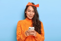 Portrait of happy pretty Asian woman with long hair, wears headband and orange sweater, holds mobile phone for chatting, downloads new application, updates software, looks positively at camera
