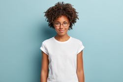 Attractive female student with curly hair, wears transparent glasses, white t shirt, stands against blue background, has calm face expression, tender smile, listens interlocutor with pleasure