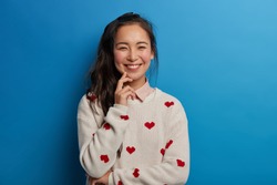 Pretty Asian woman has toothy smile, rouge cheeks, wears jumper with heart print, expresses sincere emotions, has dark hair combed in pony tail, isolated over blue background. Happiness concept