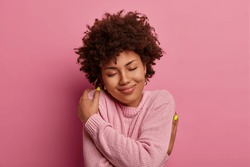 Self love and tenderness concept. Delighted gentle woman with curly hairstyle, embraces herself, hugs own body, closes eyes and wears warm soft casual jumper for cold weather, smiles cheerfully