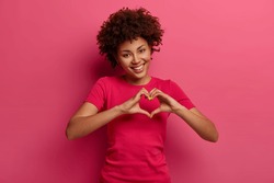 Pretty curly African American woman confesses in love, makes heart gesture, shows her true feelings, has happy expression, wears casual red t shirt, poses over pink background. Relationship concept