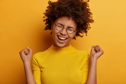 Cheerful triumphing woman achieves victory, raises clenched fists with triumph, rejoices winning prize, dressed casually, keeps eyes closed, isolated over yellow background. Celebration concept