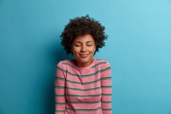 Portrait of pleasant looking dark skinned woman stands with eyes shut, being in good mood, imagines something nice, dreams about perfect vacation, wears striped sweater, isolated on blue background
